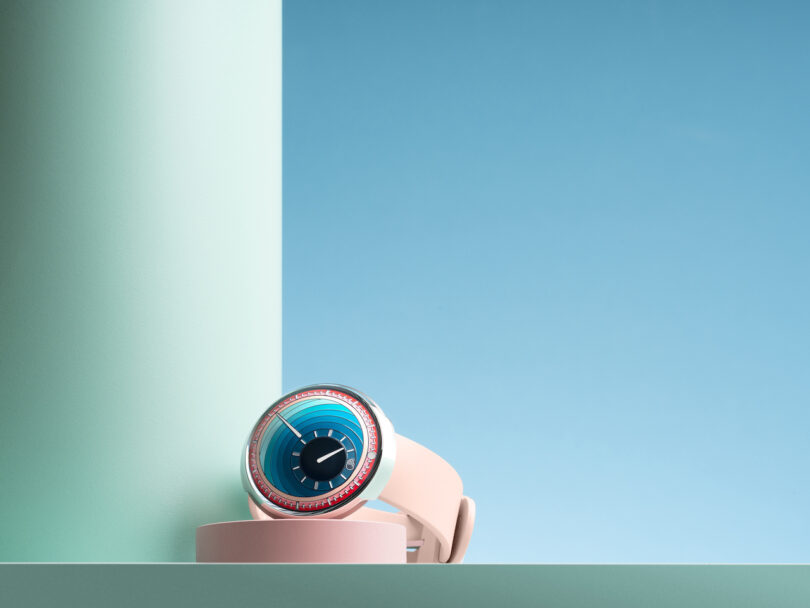 A modern wristwatch with a circular, blue and turquoise face, red detailing, and a light beige strap, displayed on a light blue background