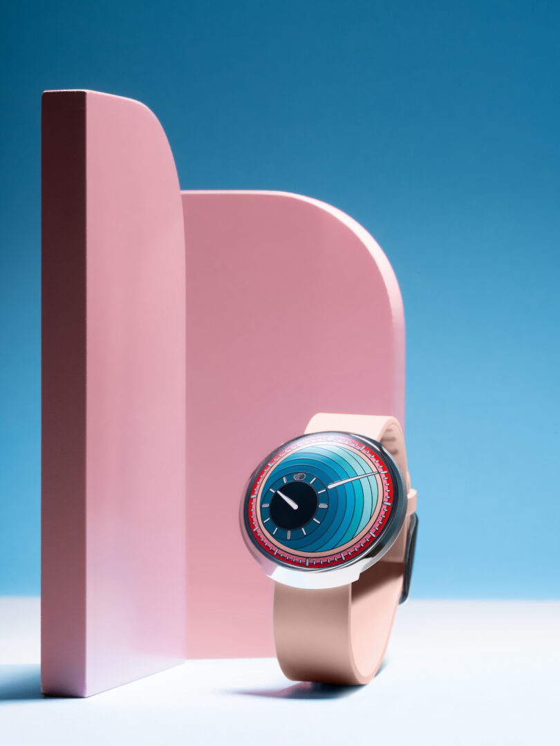 A modern wristwatch with a blue and red face and a light pink strap is displayed upright against pink geometric shapes on a blue background