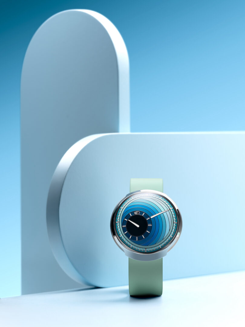 A modern wristwatch with a green strap and a circular blue and black face is displayed against a minimalist blue geometric background