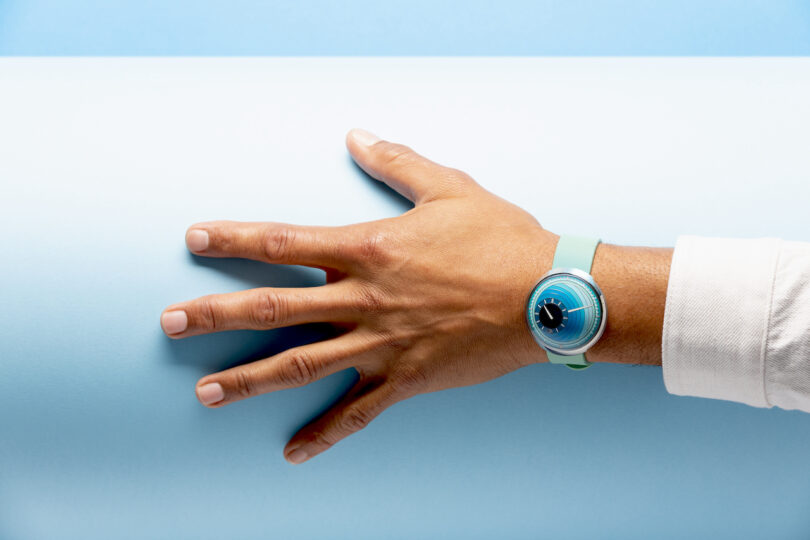A person's hand with an aqua blue wristwatch rests on a rolled sheet of light blue paper, set against a blue background