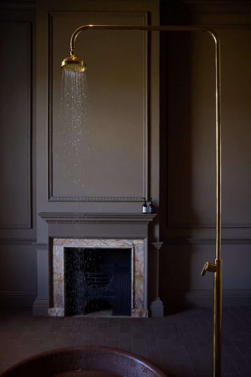 A brass shower with water running stands in front of a marble fireplace in a dimly lit room with panelled walls.