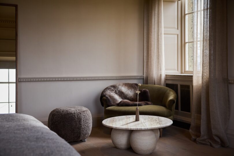 A cozy corner with an olive green armchair, a fur throw, a round pouf, a marble coffee table with a vase, and soft natural light from a large window with sheer curtains.