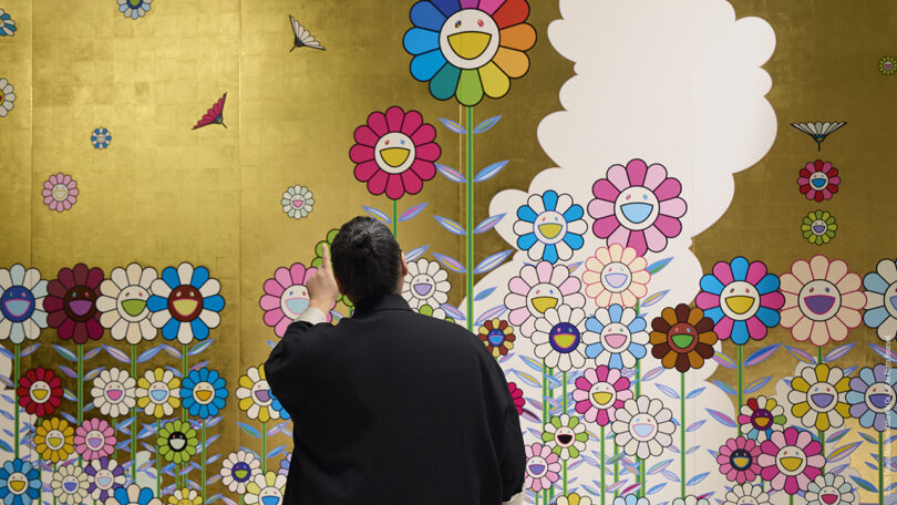 A person stands in front of a mural featuring colorful, smiling cartoon flowers and butterflies on a gold background.