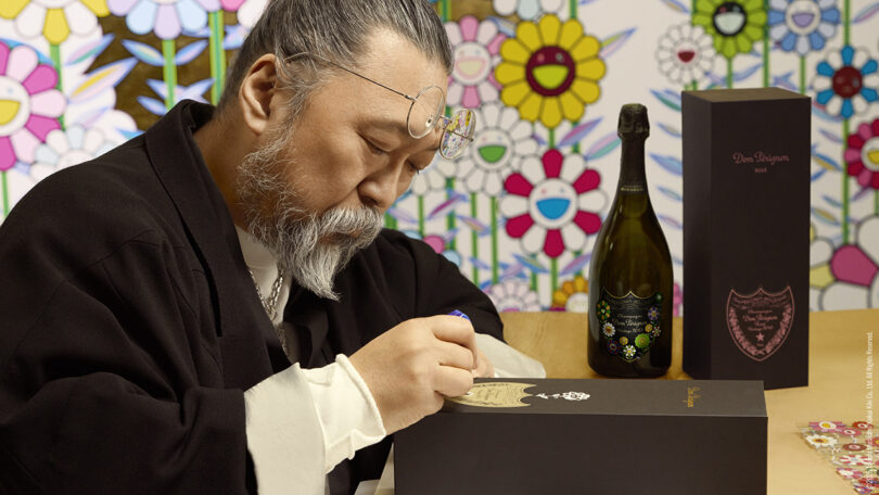 A man with gray hair and a beard signs a black box at a table, with a decorated bottle and floral-patterned backdrop behind him.