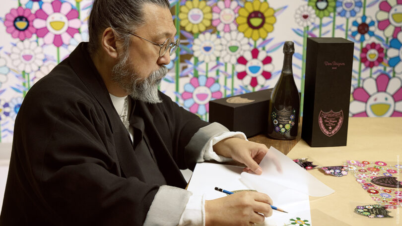 A man sits at a table drawing colorful flowers on paper with a pencil, surrounded by bottles and boxes with floral designs, against a background of cartoon-like flower art.
