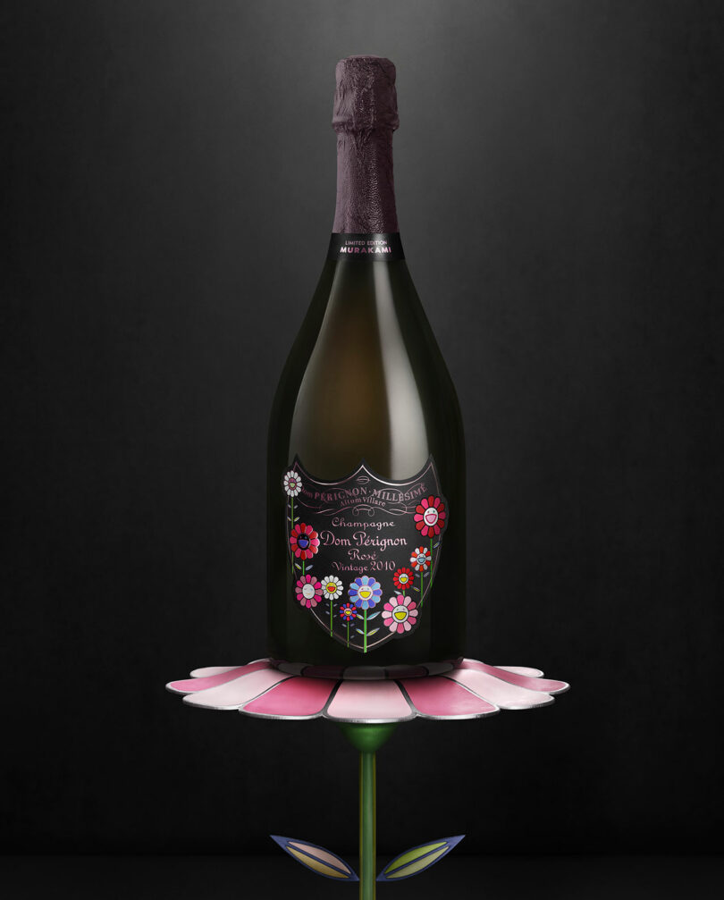 A bottle of Takashi Murakami Dom Perignon champagne with a floral-themed label is displayed on a pink and green flower-shaped stand against a dark background.