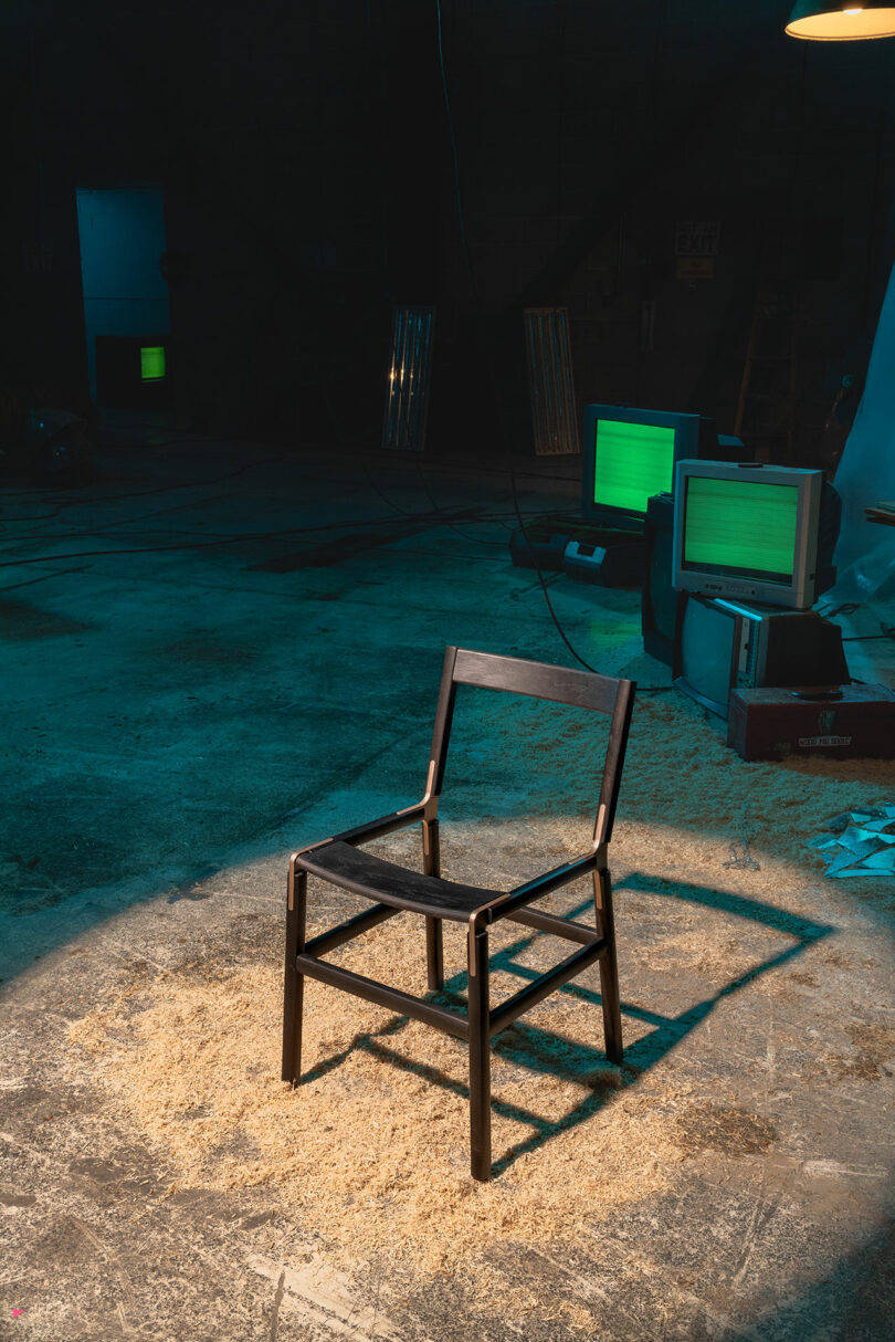A wooden chair stands on a sawdust-covered floor in a dimly lit room with old computer monitors in the background.