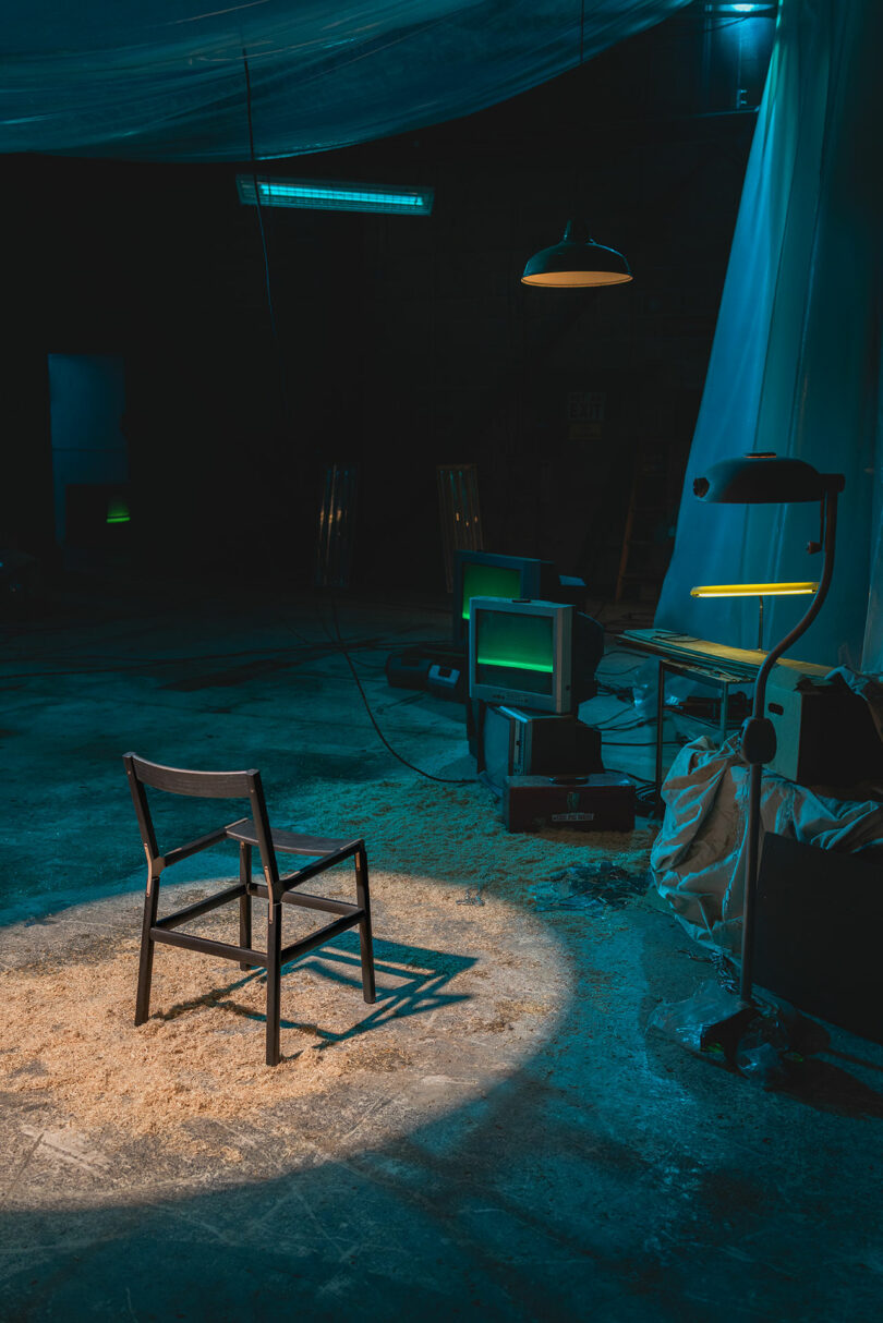 A single wooden chair is illuminated by a spotlight in a dimly lit, industrial room with scattered old electronics and plastic sheeting.