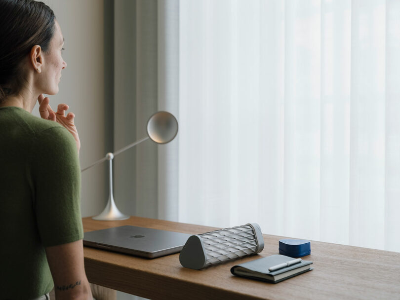 A person sits at a wooden desk with a closed laptop, a notebook, pen, a modern desk lamp, an Elio Water Bottle, Lumio book light, and small accessories, looking out a window with sheer curtains.
