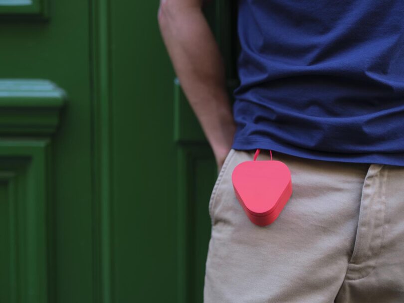 A person wearing beige pants and a navy shirt stands with a red, heart-shaped object clipped to their pocket in front of a green door.