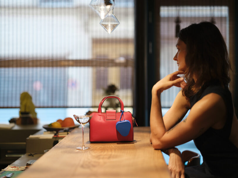 A woman sits at a wooden counter next to a pink handbag and a cocktail glass, with soft lighting and a blurred background.