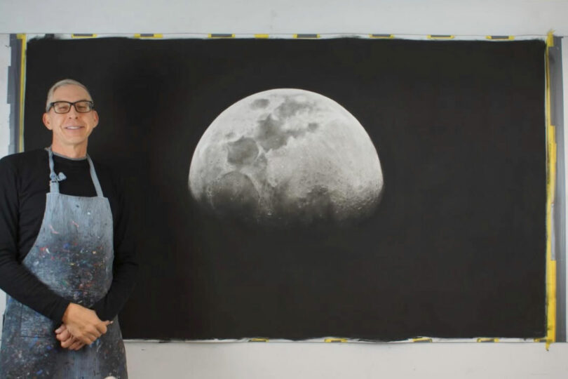 A person wearing glasses and an apron stands next to a large, detailed black-and-white painting of the moon on a wall.