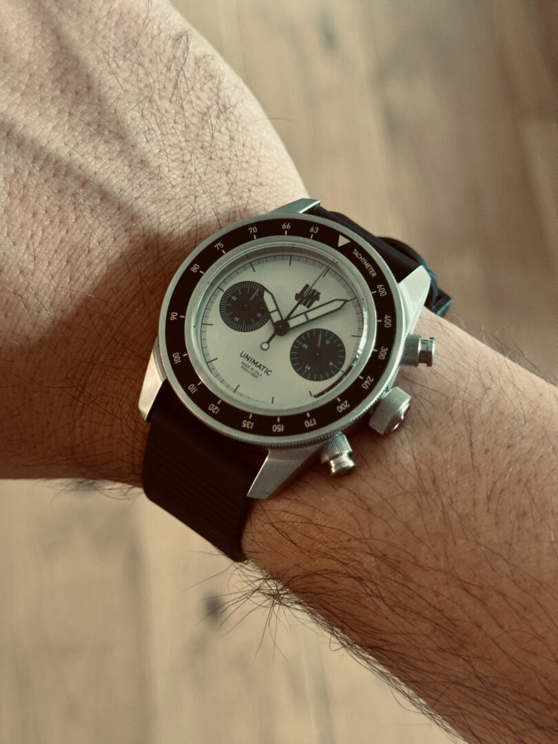 A wrist with a black and silver chronograph wristwatch displaying three subdials and a black strap, shown against a blurred wooden background.