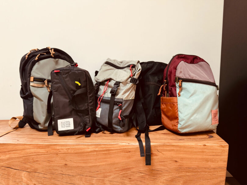 Four backpacks of different colors and styles are placed side by side on a wooden surface against a plain background.