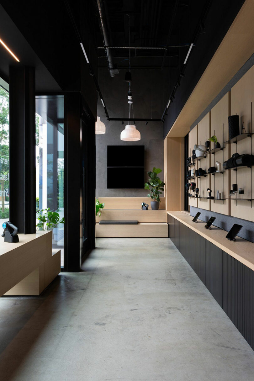 Modern retail store interior with minimalist design, featuring electronic devices displayed on wall shelves, wood accents, concrete floor, and potted plants by large windows.
