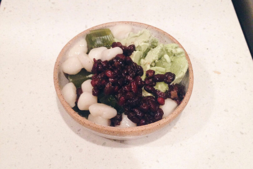 A bowl of Japanese dessert with green tea ice cream, sweet red beans, mochi, and green jelly cubes on a white countertop.