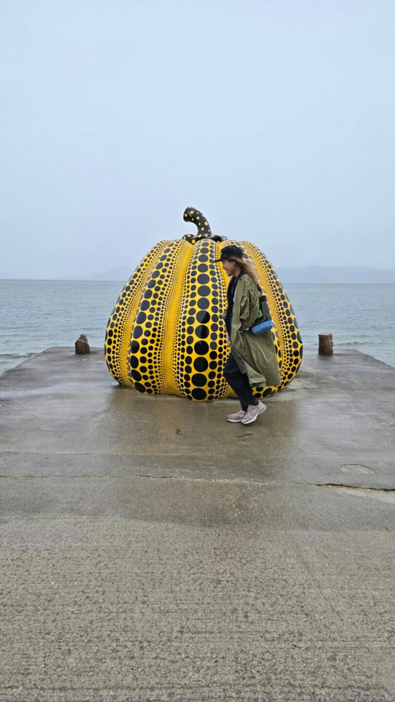 A person walks in front of a large yellow pumpkin sculpture with black dots, situated on a concrete pier by the sea under a cloudy sky.