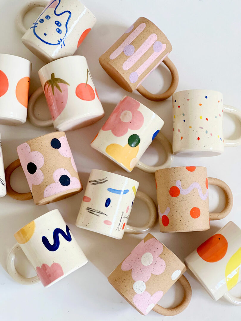 A collection of ceramic mugs with various colorful, abstract, and playful hand-painted designs arranged on a white surface.