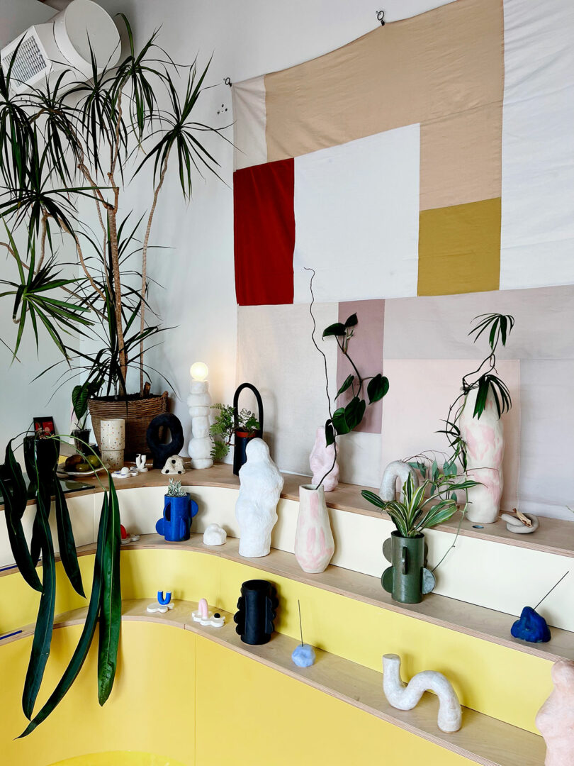 A display of abstract ceramic and clay sculptures, plants, and vases on tiered wooden shelves against a geometric wall hanging in a brightly lit room with yellow accents.