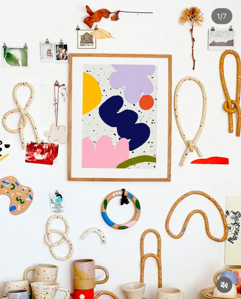 A colorful abstract art print is centered on a white wall, surrounded by ceramic loops, mugs, small photos, and decorative objects arranged in an eclectic display.