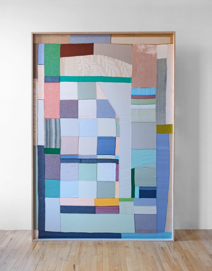 A framed abstract geometric textile artwork by Kiva Motnyk hangs on a wall, illuminated by natural light from a nearby window.