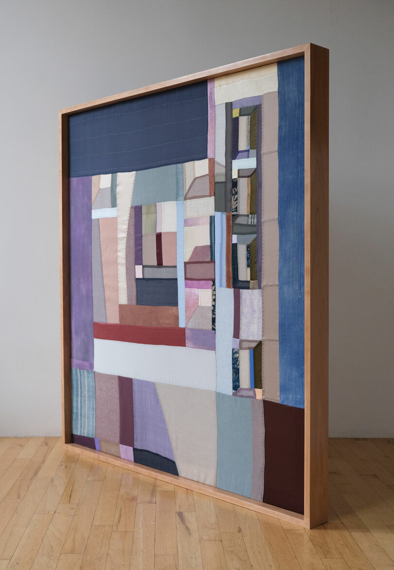 A framed abstract geometric textile artwork by Kiva Motnyk hangs on a wall, illuminated by natural light from a nearby window.