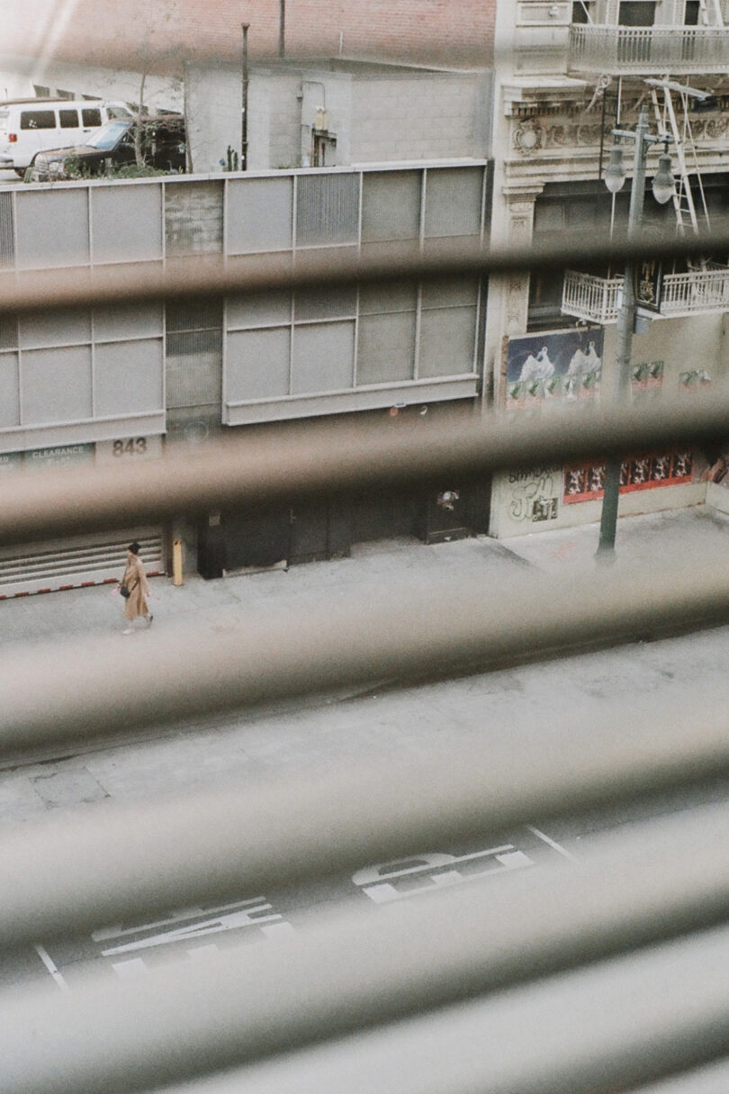 View through window blinds onto a street, showing a person in a long coat walking on the sidewalk next to a building and a parked car in the background.
