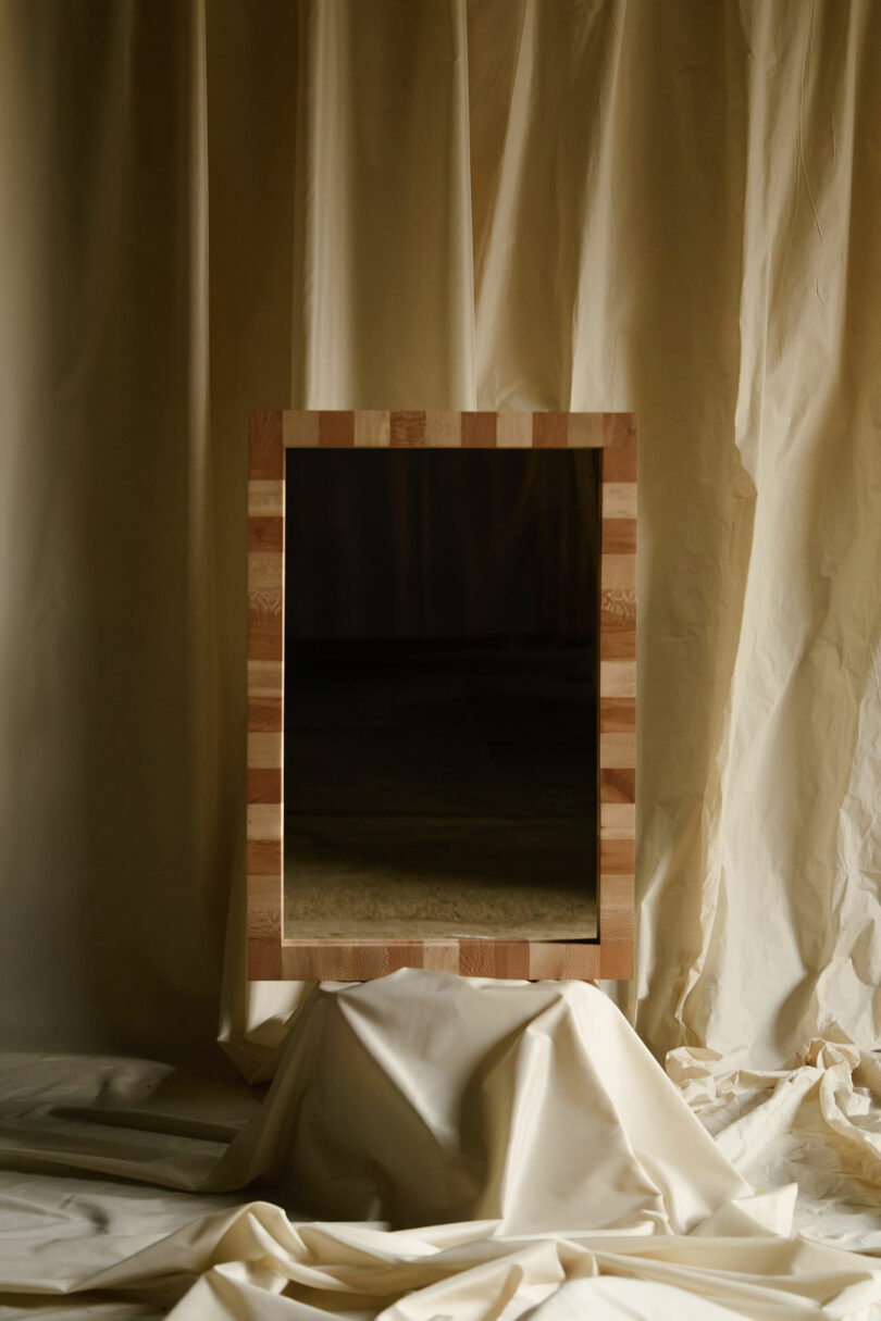 A rectangular mirror with a wooden frame is propped on draped, off-white fabric with a matching fabric backdrop.