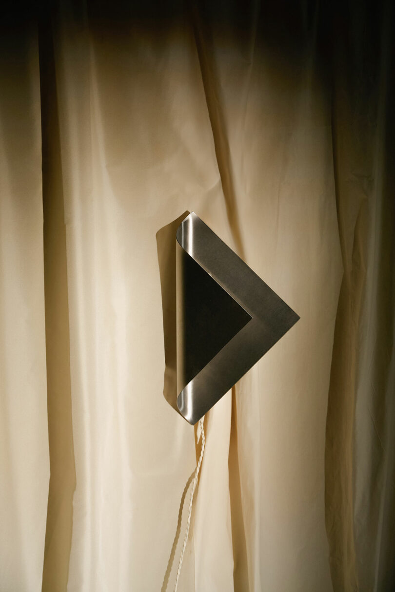 A triangular metallic object is mounted on a beige, draped curtain, with a cord hanging down from its bottom edge.