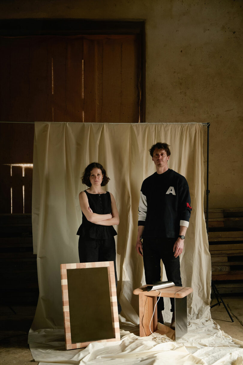 Two people stand side by side in front of a beige backdrop, with a framed mirror and a small wooden table with objects on it placed in front of them.