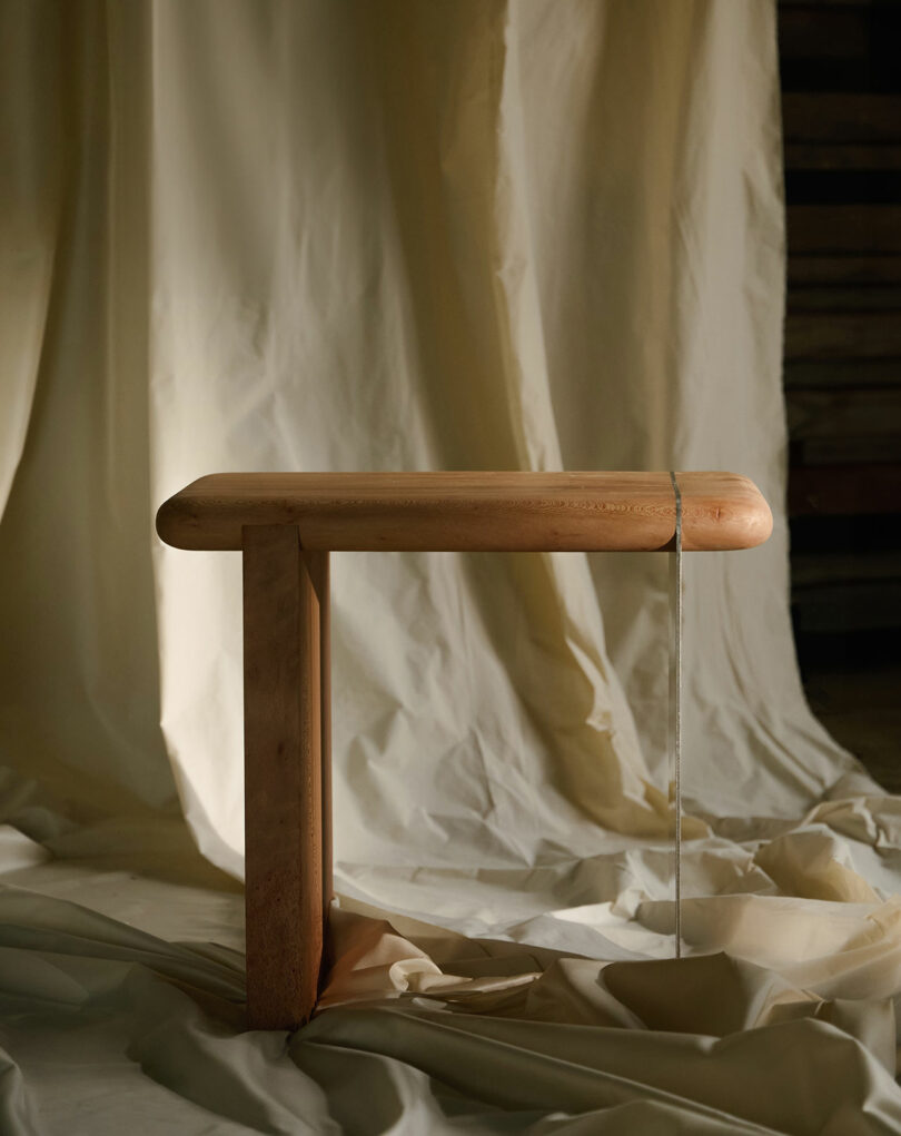 A wooden, L-shaped bench stands upright on draped cream fabric with soft, neutral lighting and a string hanging from its edge.
