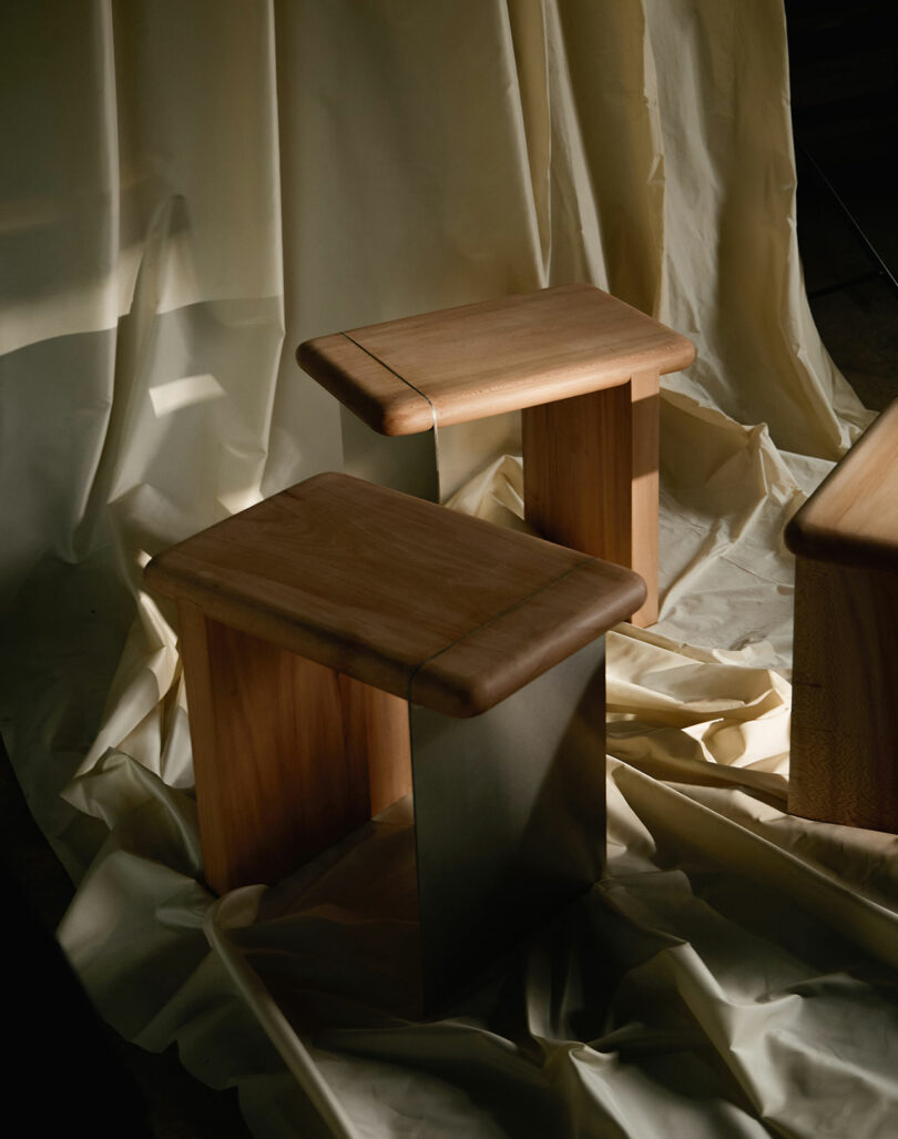 Two wooden stools with rectangular seats and legs are placed on a wrinkled cream-colored fabric backdrop, with soft light illuminating the scene.