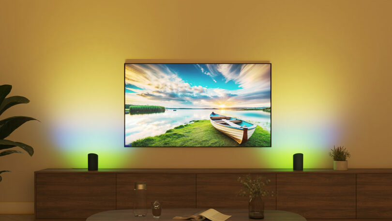 A wall-mounted TV displays a scenic image of a boat on a lake at sunset, enhanced by colorful ambient lighting behind the screen, showcasing the beauty of technology alongside two speakers on a wooden cabinet below.