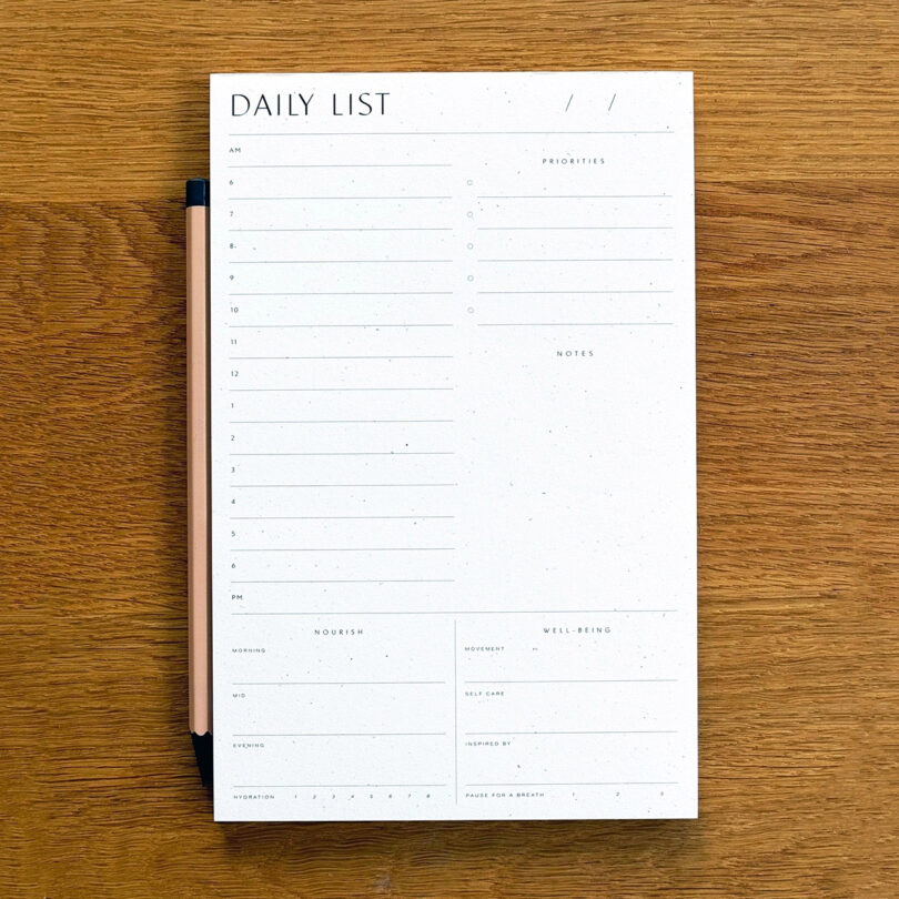 A blank daily planner sheet with sections for hourly scheduling, priorities, notes, morning routines, well-being, and a pen placed along the left edge, all on a wooden surface.