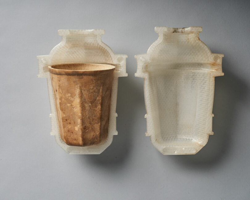 A brown paper cup placed in a translucent, textured, two-part plastic mold, with one half of the mold open and empty beside it on a gray surface.