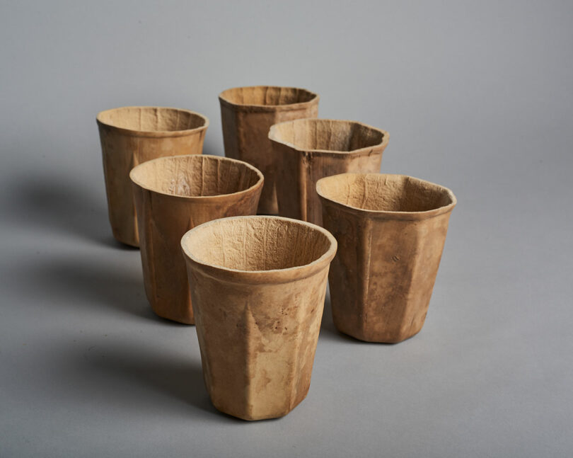 Six brown, textured cups made from a natural-looking material are arranged on a plain gray surface.
