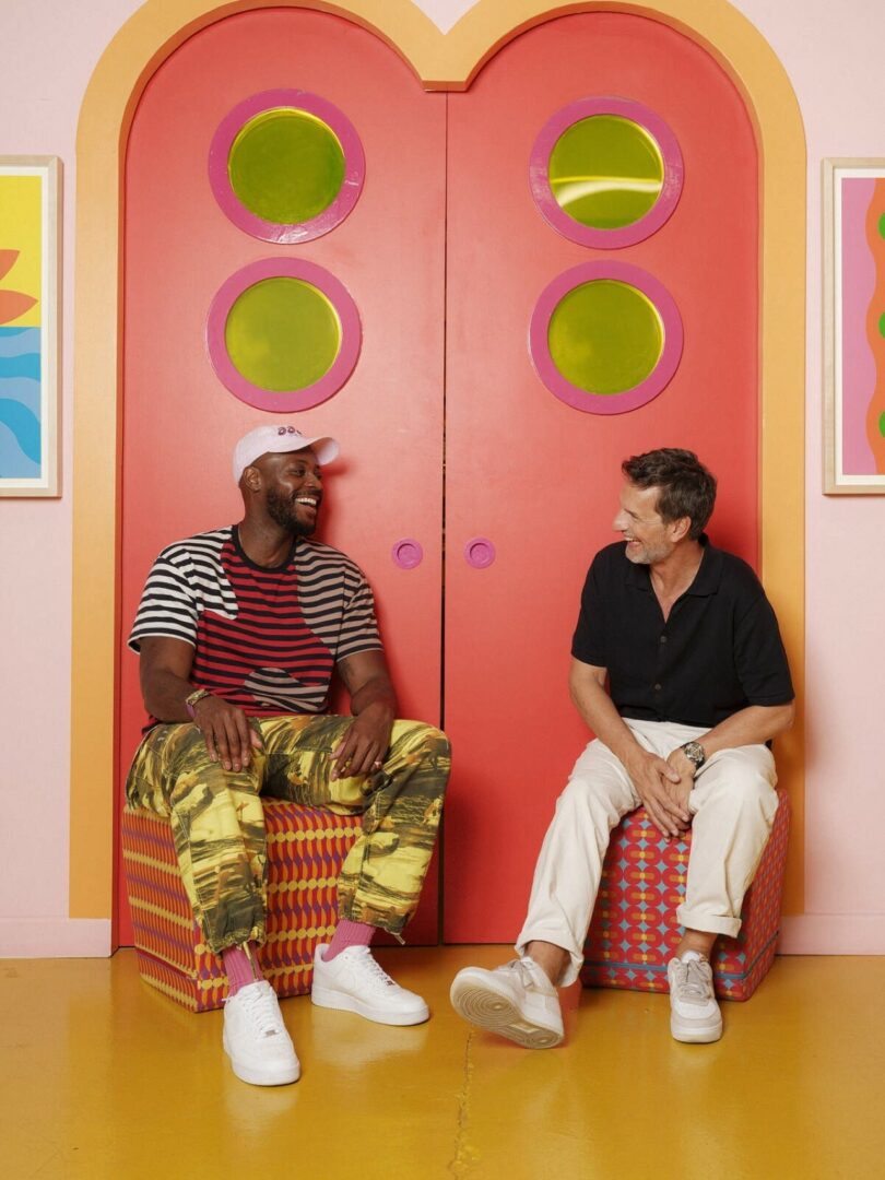 Two men sit on patterned chairs in front of large, pink double doors with circular windows, smiling and talking. The room has colorful decor and art on the walls