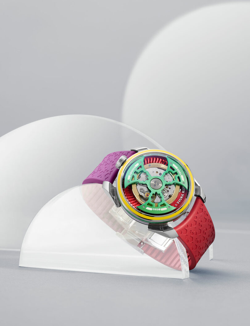 A colorful wristwatch with a red and purple band, a multi-colored bezel, and visible internal mechanics, displayed on a clear, curved stand against a light gray background