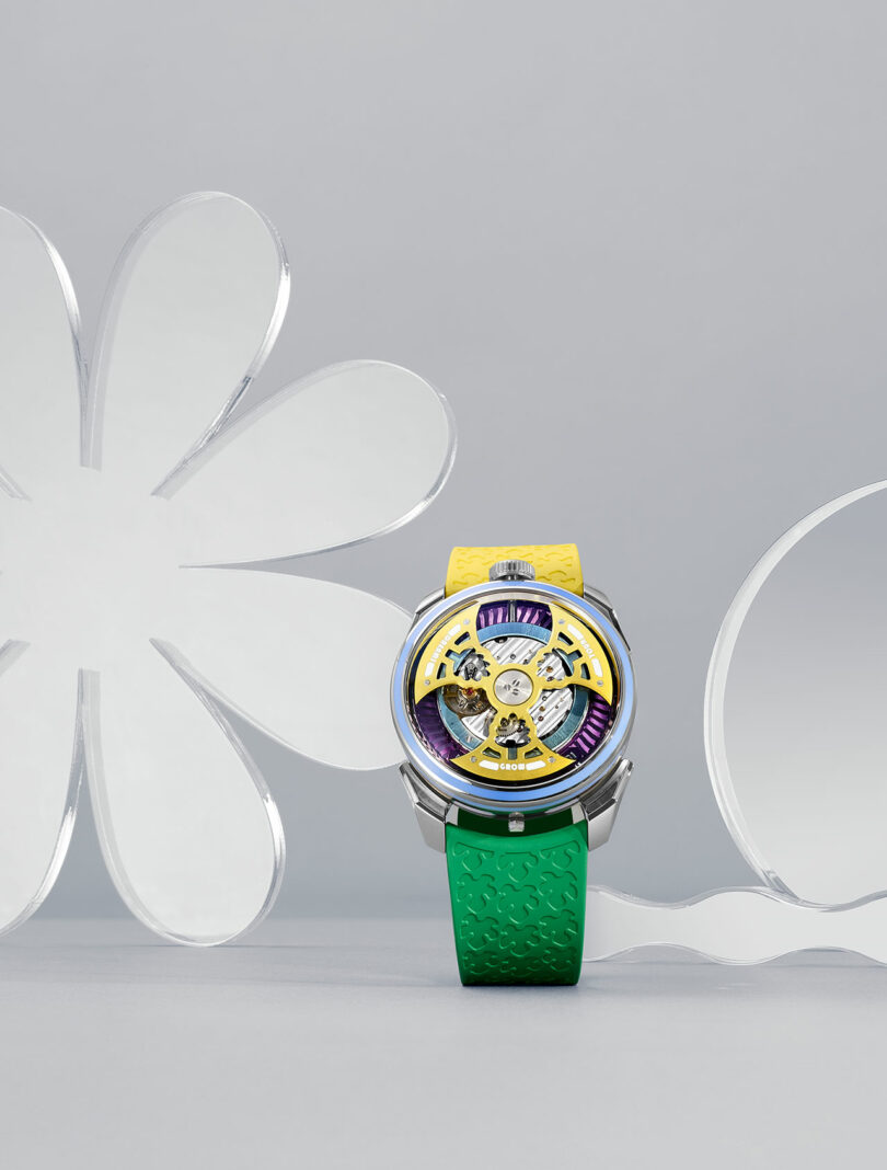 A wristwatch with a yellow and green band, yellow and silver face, and visible mechanical parts, displayed against a gray background with clear, flower-shaped acrylic decorations