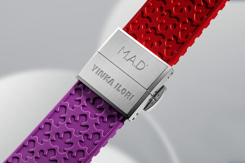 Close-up of a watch clasp engraved with "M.A.D." and "YINKA ILORI," attached to textured red and purple rubber straps