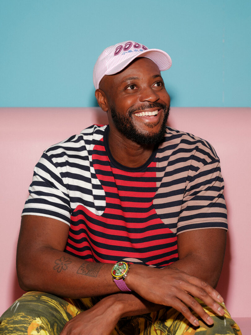 A man wearing a striped shirt, patterned pants, a pink cap, and a colorful watch sits on a pink bench against a blue wall, smiling and looking to the side