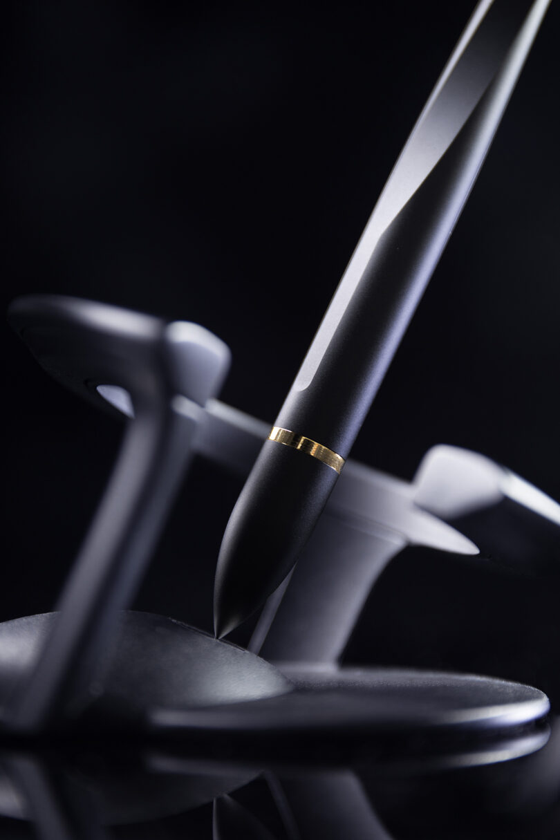 A close-up of a black Novium Hoverpen with a gold ring detail resting on a modern black pen holder, set against a dark background.