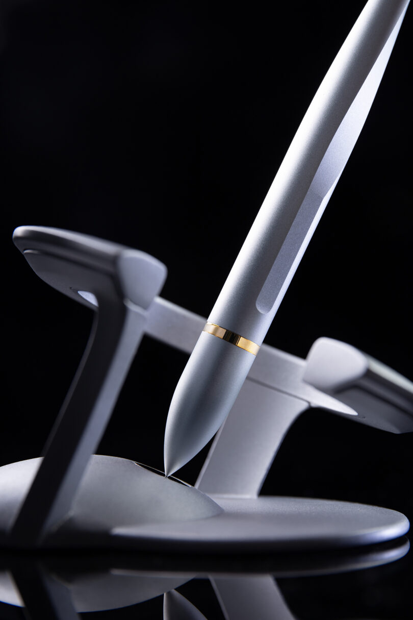 A Novium Hoverpen, silver with a gold band, is placed vertically on a round, metallic stand against a black background.