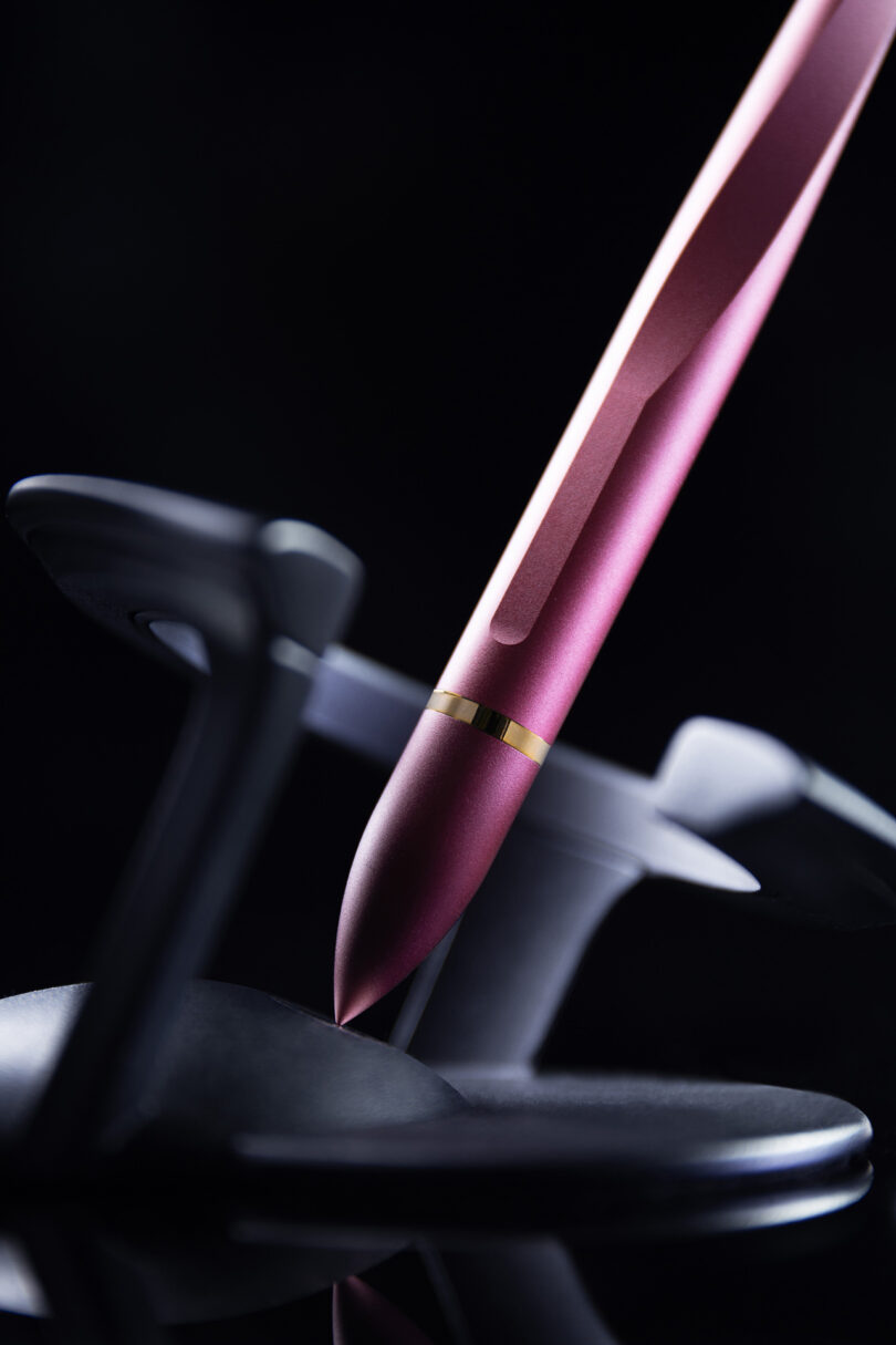 A pink metallic Novium Hoverpen with a gold band rests upright in a gray stand against a dark background.