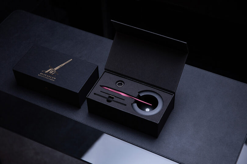 A luxury Novium Hoverpen fountain pen set with a red pen, ink bottle, and accessories displayed in an open black presentation box next to a closed box on a dark surface.
