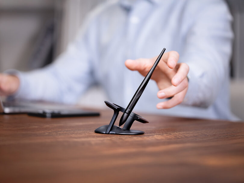 A person reaches towards a black Novium Hoverpen balanced upright in a modern pen holder on a wooden desk, with a laptop nearby.