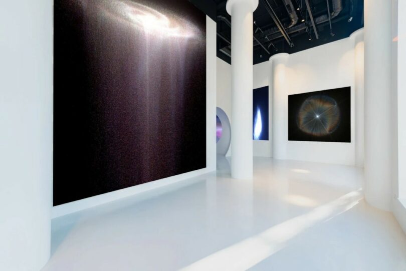 A modern art gallery with white walls and floors, featuring large abstract artworks in dark colors and circular patterns displayed on the walls