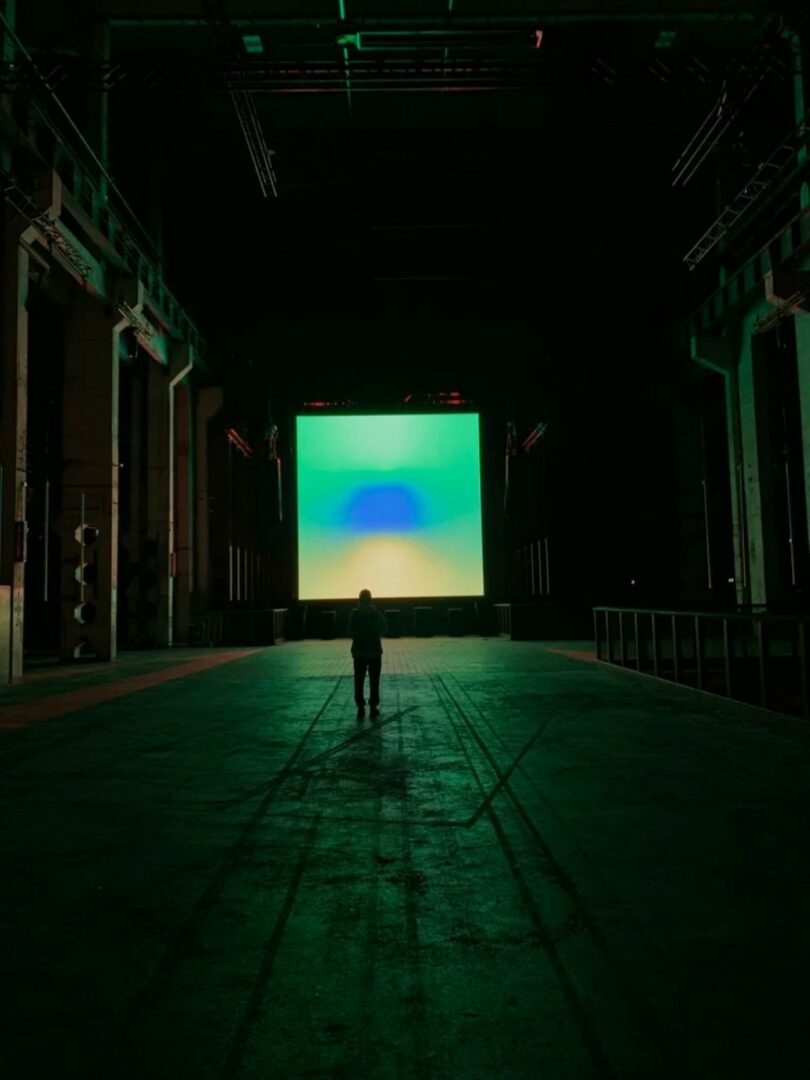 A person stands alone in a large, dimly lit industrial space facing a bright, abstract square screen displaying green and blue colors