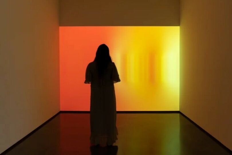 A person in a long dress stands in front of a brightly lit, gradient wall transitioning from red to yellow in a dim, minimalist room