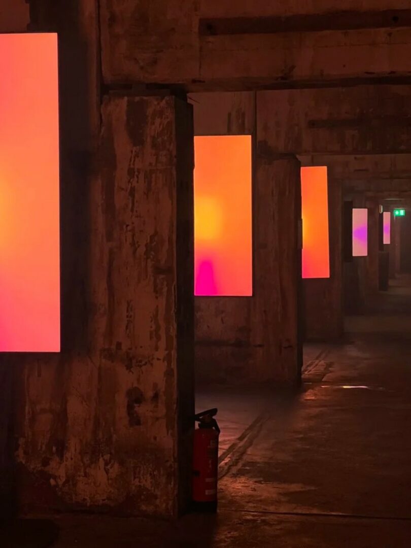 Large rectangular panels display pink and orange gradients on worn concrete pillars in a dimly lit industrial corridor; a red fire extinguisher sits on the floor